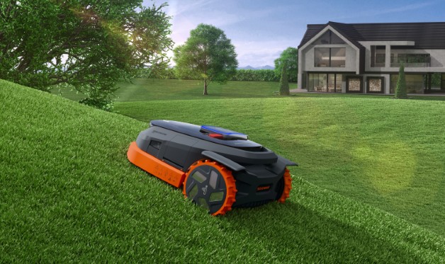Bildet viser en Segway Navimow robotgressklipper som klipper gresset på en bratt helling foran et moderne hus. Gressklipperen er utstyrt med store hjul og et robust design som gjør den i stand til å takle skråninger effektivt. Den autonome gressklipperen navigerer selvstendig i terrenget, og viser hvordan Navimow kan håndtere utfordrende landskap samtidig som den gir en jevn og presis klipp. Foto