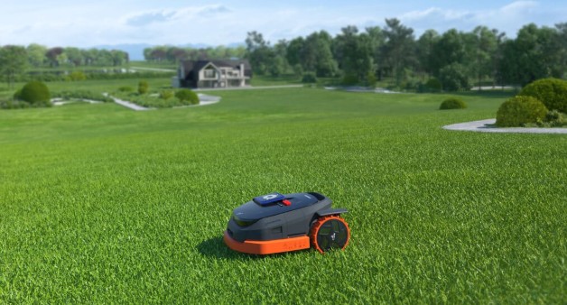Bildet viser en robotgressklipper som arbeider på en stor, velstelt plen i et åpent landskap. I bakgrunnen kan man se en moderne bolig omgitt av grøntområder. Gressklipperen er designet for å håndtere store plener og navigerer selvstendig med presisjon, noe som sikrer jevn klipping over hele området. Foto